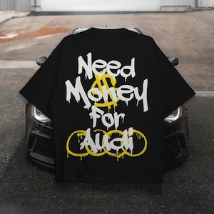 May include: Black t-shirt with the text "Need Money For Audi" in white dripping font, with a yellow dollar sign and Audi logo. The shirt is displayed over the hood of a black car.