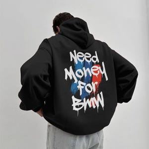 May include: Black hooded sweatshirt with the text "Need Money For BMW" in white, with blue and red paint strokes. The sweatshirt has a drawstring hood and long sleeves. The back of the sweatshirt is visible.