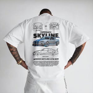 May include: White short-sleeve t-shirt featuring a graphic of a silver and blue Nissan Skyline GTR R34 car. The design includes blueprints and text that reads "NISSAN SKYLINE GTR R34".