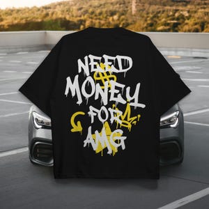 以下が含まれることがあります： 黒いTシャツ。白と黄色のグラフィティ風のテキストで「NEED MONEY FOR AMG」と書かれています。デザインにはドル記号と王冠が含まれています。Tシャツはシルバーの車の前に表示されています。
