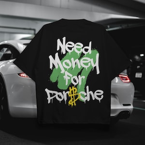 Pode incluir: Camiseta preta com a frase "Need Money For Porsche" em letras brancas e verdes em estilo grafite. O símbolo do dólar em "Porsche" é amarelo. A camiseta é exibida em frente a um carro branco.