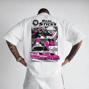 Könnte beinhalten: Weißes Kurzarm-T-Shirt mit einem grafischen Design auf dem Rücken. Das Design zeigt einen Comicstrip mit rosa und schwarzen Illustrationen von Autos und Fahrern sowie den Text "REAL CHICKS DRIVE STICKS".
