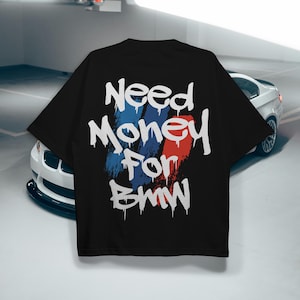 May include: Black t-shirt with the text "Need Money For BMW" in white, with blue and red brushstroke accents. The shirt is displayed in front of a white BMW car.