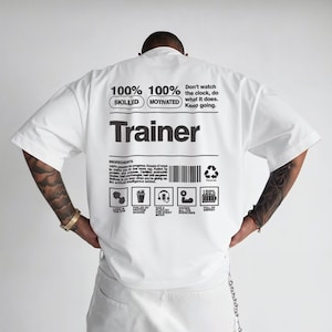 May include: White short-sleeved t-shirt with the word "Trainer" printed in black. The back of the shirt features text including "100% Skilled, 100% Motivated" and a barcode. The shirt has a casual, athletic style.