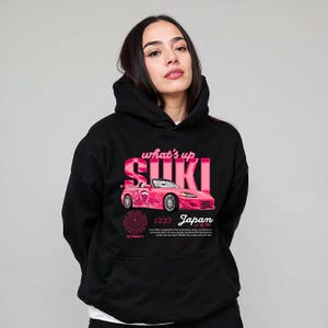 May include: Black hoodie featuring a pink graphic of a convertible car with the text "what's up SUKI Japan". The design includes a circular emblem and additional text at the bottom. The hoodie has a front pocket and a hood.