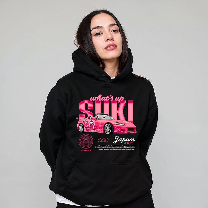 May include: Black hoodie featuring a pink graphic of a convertible car with the text "what's up SUKI Japan". The design includes a circular emblem and additional text at the bottom. The hoodie has a front pocket and a hood.