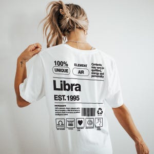 May include: White t-shirt with a Libra zodiac design. The shirt features the word "Libra" in bold black letters, along with the text "EST. 1995" and other graphics. The back of the shirt also has the words "100% UNIQUE ELEMENT AIR".