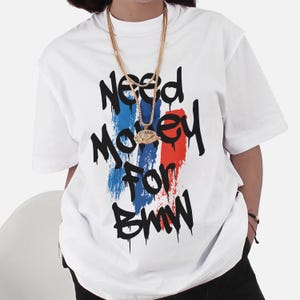 May include: White t-shirt with the text "Need Money For BMW" in black dripping font. The text is over a vertical stripe design in blue, red, and white. A gold chain necklace is worn over the shirt.