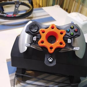 May include: A black and silver gaming steering wheel with an orange star-shaped adapter. The steering wheel has buttons labeled "LSB", "RSB", "X", "Y", "A", and "B".