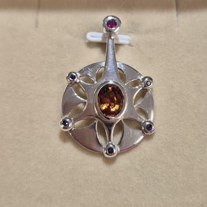 May include: A silver pendant with an oval, amber-colored gemstone at the center. The pendant has a circular design with eight points, each adorned with a small, dark blue gemstone. A smaller, round, red gemstone sits at the top.