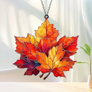 May include: A stained glass suncatcher shaped like a cluster of maple leaves in vibrant shades of red, orange, and yellow. The leaves feature dark leaded outlines and are suspended from a thin metal chain, creating a translucent autumn display.