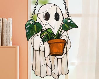 May include: A stained glass ghost holding a potted plant, suspended by a chain. The ghost is white with black eyes and a stitched appearance. The plant has large green leaves and sits in an orange pot. The background is a window with white curtains.