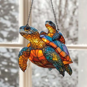 May include: A stained glass turtle suncatcher with a baby turtle riding on its back. The turtles are in shades of blue, orange, and green, with a chain for hanging. The suncatcher is designed to catch the light.
