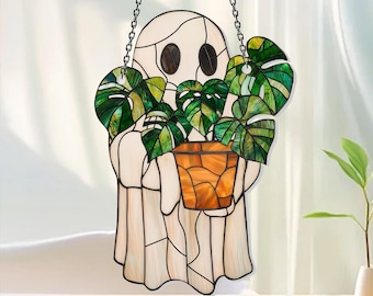 May include: A stained glass suncatcher shaped like a ghost holding a potted monstera plant. The ghost is off-white with dark oval eyes, while the plant features vibrant green leaves and the pot is constructed from various shades of orange glass.