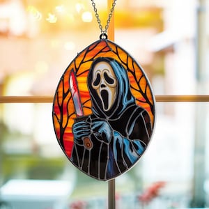 May include: An oval stained glass suncatcher featuring a hooded figure with a white ghost mask holding a silver knife with red accents. The background depicts black tree silhouettes against an orange and yellow sunset sky, suspended by a metal chain.
