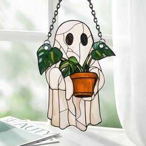 May include: A stained glass window hanging with a white ghost holding a potted plant with green leaves. The ghost has two black eyes and a white background.