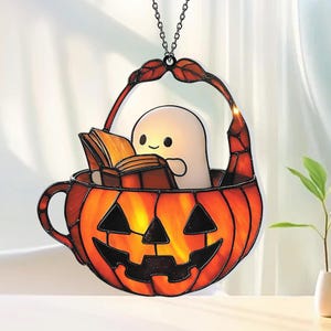 Może przedstawiać: Dekoracja witrażowa o tematyce Halloween. Przedstawia uśmiechniętego ducha czytającego książkę w pomarańczowym koszu w kształcie dyni. Kosz ma uchwyt i jest zawieszony na łańcuchu. Duch i dynia mają czarne akcenty.