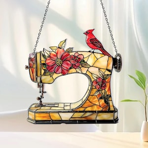 May include: A stained glass suncatcher shaped like a sewing machine, featuring a red cardinal perched on top. The design includes yellow and amber glass panels with red floral accents and green leaves, suspended by a thin metal chain for display.