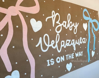 May include: Brown banner with pink and blue bows, white hearts, and the text "Baby Velazquez IS ON THE WAY" in white script. White dots are scattered across the banner. The banner is likely for a baby shower.