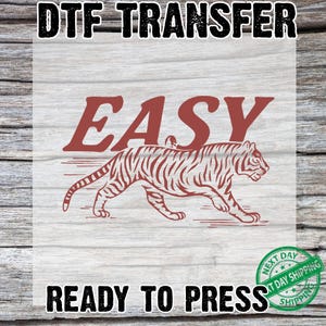 May include: DTF transfer with the word "EASY" above a red tiger illustration. The words "READY TO PRESS" are below the tiger. The background is a light wood grain pattern.