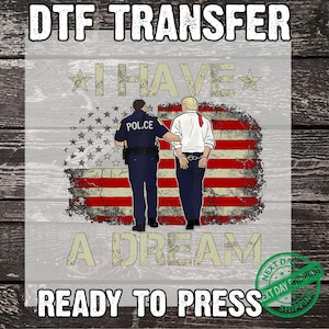 May include: DTF transfer graphic featuring a police officer escorting a man in handcuffs in front of a distressed American flag. Text includes "I HAVE A DREAM" and "READY TO PRESS". The design is on a light gray background.
