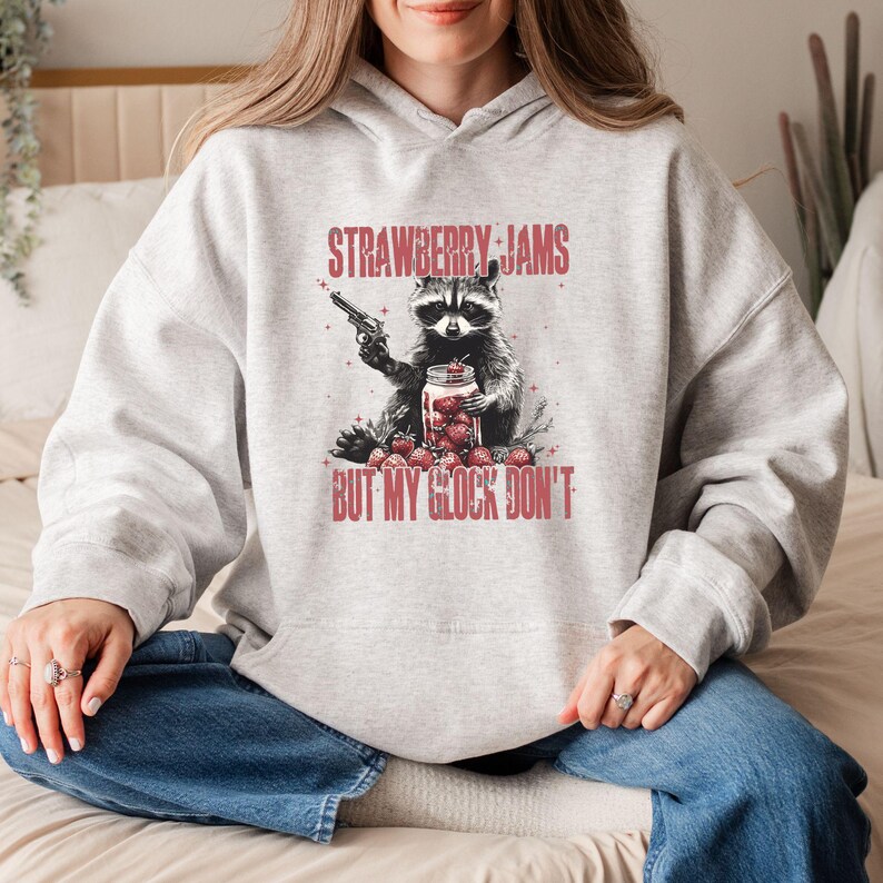 Pu&ograve; includere: Felpa con cappuccio grigio chiaro con una grafica di un procione che tiene una pistola e un barattolo di fragole. Il testo rosso dice "STRAWBERRY JAMS BUT MY GLOCK DON'T". Il procione &egrave; bianco e nero, le fragole sono rosse.