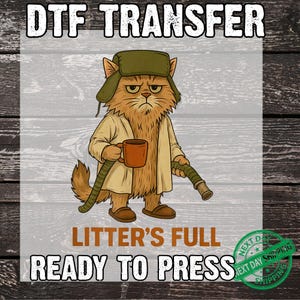 May include: A DTF transfer graphic featuring a grumpy orange cat wearing a green trapper hat, a beige coat, and slippers. The cat holds a brown mug and a green hose. Text reads DTF TRANSFER, LITTER'S FULL, and READY TO PRESS in bold lettering.