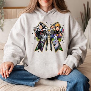 May include: A light gray hoodie featuring a large bow design with the word "Villains" and illustrations of Disney villains. The bow has black ribbons and is decorated with spiderwebs and stars. The hoodie is made of a soft material.