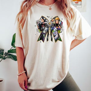 May include: Cream-colored t-shirt featuring a bow design with the word "Villains" and illustrations of cartoon characters. The bow has a gold border and is decorated with bats and spiderwebs. The shirt is a comfortable, casual style.