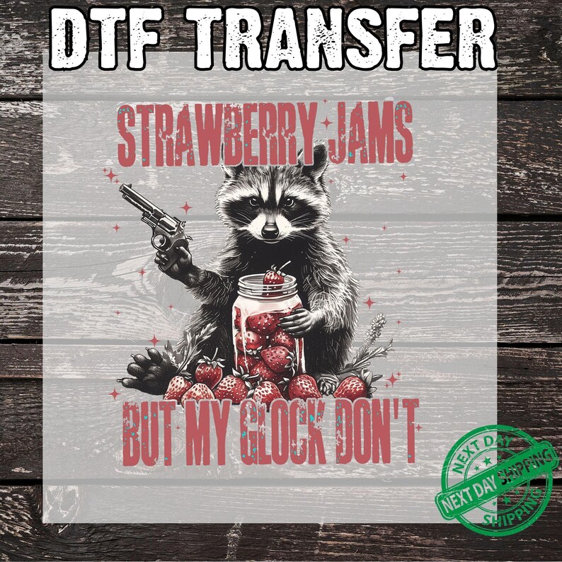 Pu&ograve; includere: Trasferimento DTF con un'illustrazione di un procione che tiene una pistola e un barattolo di fragole. Il testo recita "STRAWBERRY JAMS BUT MY GLOCK DON'T". Il design utilizza toni rossi, bianchi e grigi, con uno stile rustico.