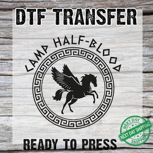 May include: A black DTF transfer featuring a silhouette of a winged horse inside a circular Greek key design. The text "LAMP HALF-BLOOD" and "READY TO PRESS" are also included. The image is on a light wood grain background.