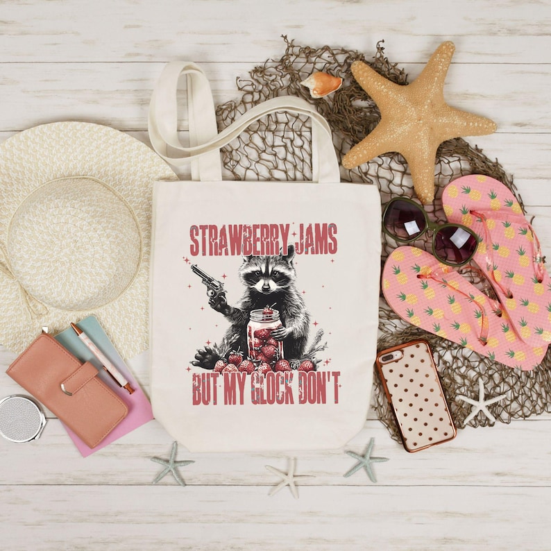 Pu&ograve; includere: Borsa tote color crema con la scritta "STRAWBERRY JAMS BUT MY GLOCK DON'T" e una grafica di un procione che tiene una pistola e un barattolo di fragole. Accessori da spiaggia, tra cui un cappello e occhiali da sole.