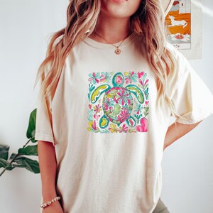 以下が含まれることがあります： クリーム色のTシャツ。カラフルな花の亀のデザインが特徴です。亀はピンク、緑、黄色の花で飾られ、ターコイズとピンクの花のアクセントに囲まれています。カジュアルで快適なスタイルです。