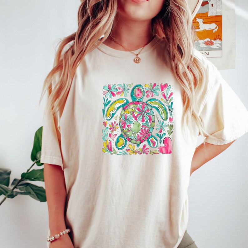 以下が含まれることがあります： クリーム色のTシャツ。カラフルな花の亀のデザインが特徴です。亀はピンク、緑、黄色の花で飾られ、ターコイズとピンクの花のアクセントに囲まれています。カジュアルで快適なスタイルです。