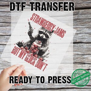 Pu&ograve; includere: Trasferimento DTF con un procione che tiene in mano una pistola e un barattolo di fragole. Il testo recita "STRAWBERRY JAMS BUT MY GLOCK DON'T" e "READY TO PRESS". Il design &egrave; in rosso e nero.