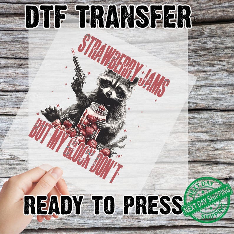 Pu&ograve; includere: Trasferimento DTF con un procione che tiene in mano una pistola e un barattolo di fragole. Il testo recita "STRAWBERRY JAMS BUT MY GLOCK DON'T" e "READY TO PRESS". Il design &egrave; in rosso e nero.