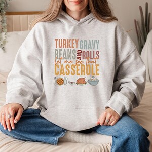 May include: A light gray hoodie with the text "TURKEY GRAVY BEANS AND ROLLS let me see that CASSEROLE" in various colors. The design includes illustrations of a pie, gravy, a turkey, and a casserole dish.