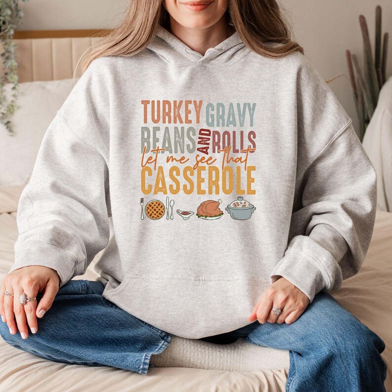 May include: A light gray hoodie with the text "TURKEY GRAVY BEANS AND ROLLS let me see that CASSEROLE" in various colors. The design includes illustrations of a pie, gravy, a turkey, and a casserole dish.