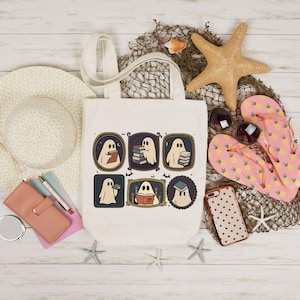 May include: Cream tote bag with six framed illustrations of cartoon ghosts reading books. The bag is surrounded by beach-themed accessories, including a straw hat, starfish, sunglasses, and pink pineapple-print flip-flops.