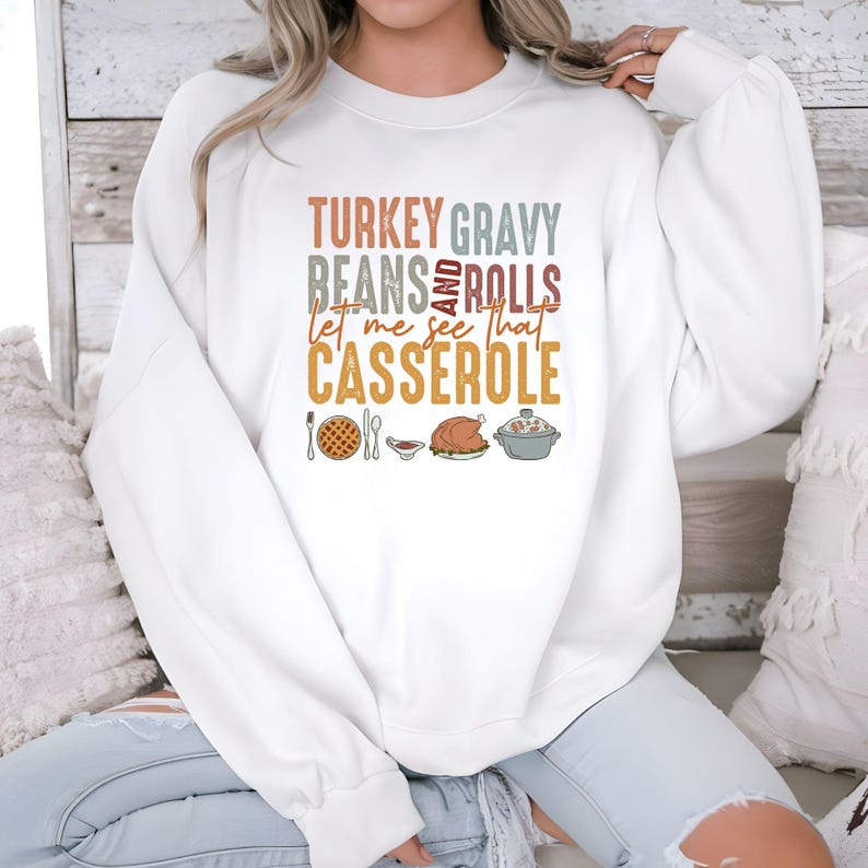 May include: White sweatshirt with the text "TURKEY GRAVY BEANS AND ROLLS let me see that CASSEROLE" in various colors. Below the text are illustrations of Thanksgiving food items, including a pie, gravy, turkey, and casserole.