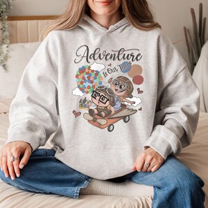 Puede incluir: Sudadera con capucha gris claro con una ilustraci&oacute;n de dibujos animados y el texto "Adventure Is Out There". El dise&ntilde;o muestra dos personajes con equipo de aviador en un avi&oacute;n, con globos y una casa. El dise&ntilde;o incluye globos de colores y corazones.