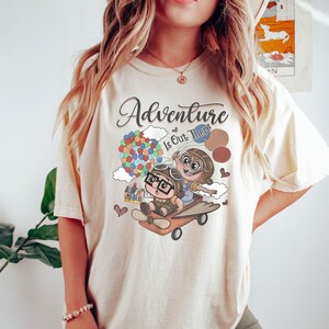 Puede incluir: Camiseta color crema con la frase "Adventure Is Out There" y un dibujo animado. El dibujo muestra dos personajes con cascos de aviador en un avi&oacute;n de madera, con globos y una casa. Peque&ntilde;os corazones est&aacute;n dispersos.