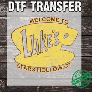 以下が含まれることがあります： 「WELCOME TO Luke's STARS HOLLOW, CT」のテキストが入った、黄色と茶色のデザインのDTF TRANSFERグラフィック。デザインはグレーと茶色の木製の背景にあります。右下には緑色の「NEXT DAY SHIPPING」スタンプがあります。