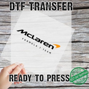 May include: A DTF (Direct to Film) transfer sheet featuring the McLaren Formula 1 Team logo in black and orange. The text "READY TO PRESS" is displayed below the logo. The image is on a wooden background.