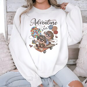 Puede incluir: Sudadera blanca con la frase "Adventure Is Out There" y un dibujo animado de dos personajes en un veh&iacute;culo volador, con un grupo de globos de colores. El dise&ntilde;o incluye corazones y una peque&ntilde;a casa.