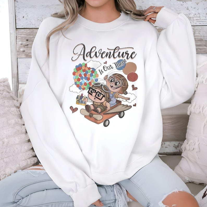 Puede incluir: Sudadera blanca con la frase "Adventure Is Out There" y un dibujo animado de dos personajes en un veh&iacute;culo volador, con un grupo de globos de colores. El dise&ntilde;o incluye corazones y una peque&ntilde;a casa.