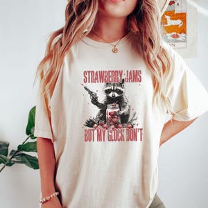 Pu&ograve; includere: T-shirt color crema con un procione che tiene in mano una pistola e un barattolo di fragole. Il testo "STRAWBERRY JAMS" e "BUT MY GLOCK DON'T" &egrave; stampato sopra e sotto l'illustrazione del procione in rosso.