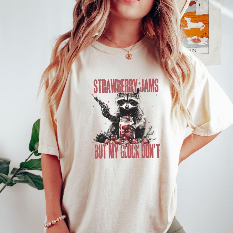 Pu&ograve; includere: T-shirt color crema con un procione che tiene in mano una pistola e un barattolo di fragole. Il testo "STRAWBERRY JAMS" e "BUT MY GLOCK DON'T" &egrave; stampato sopra e sotto l'illustrazione del procione in rosso.