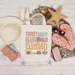 May include: Cream-colored tote bag with the text "TURKEY GRAVY BEANS AND ROLLS let me see that CASSEROLE" in various colors. The bag is surrounded by a straw hat, sunglasses, starfish, and pink pineapple-print flip-flops.