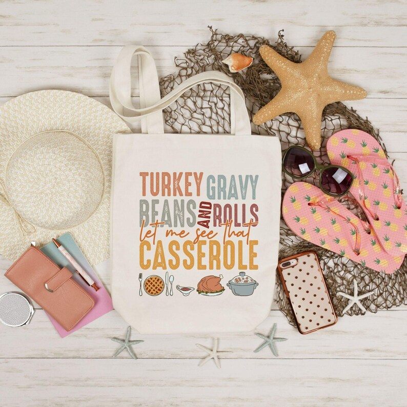 May include: Cream-colored tote bag with the text "TURKEY GRAVY BEANS AND ROLLS let me see that CASSEROLE" in various colors. The bag is surrounded by a straw hat, sunglasses, starfish, and pink pineapple-print flip-flops.